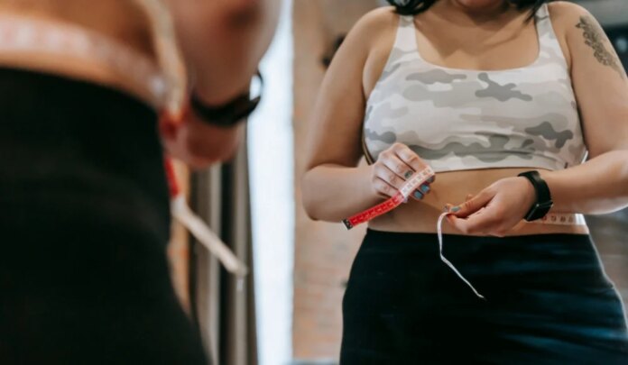 Uma pessoa veste top esportivo estampado em tons de cinza e calça preta enquanto mede a circunferência da cintura com uma fita métrica diante de um espelho. A imagem mostra apenas o tronco e as mãos, sem exibir o rosto completo. No reflexo do espelho, a fita aparece posicionada ao redor do abdômen. A pessoa usa uma pulseira ou relógio inteligente no pulso e tem uma tatuagem visível no braço direito. O ambiente interno tem iluminação suave e o fundo desfocado destaca a medição da cintura como foco principal da cena.